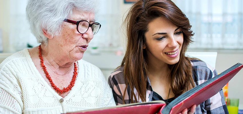 Senior woman looking at scrapbook with younger woman.