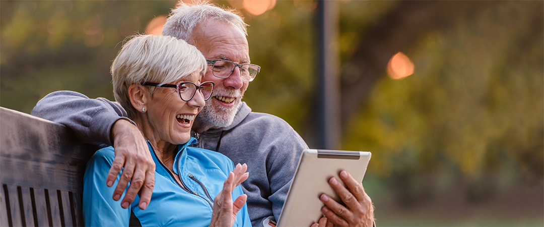 Two seniors at a park on a park bench looking at a lap top together.