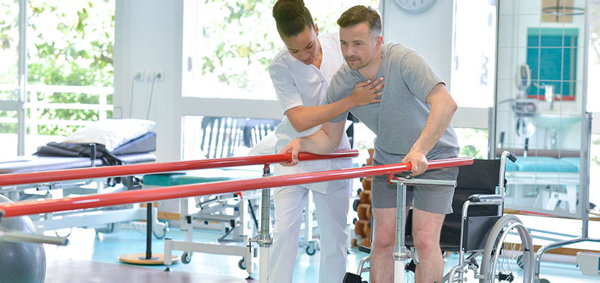 Therapist helping man with the parallel bars in a rehab gym.