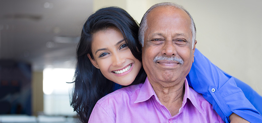 Senior man in pink shirt with younger woman in blue shirt smiling behind him.