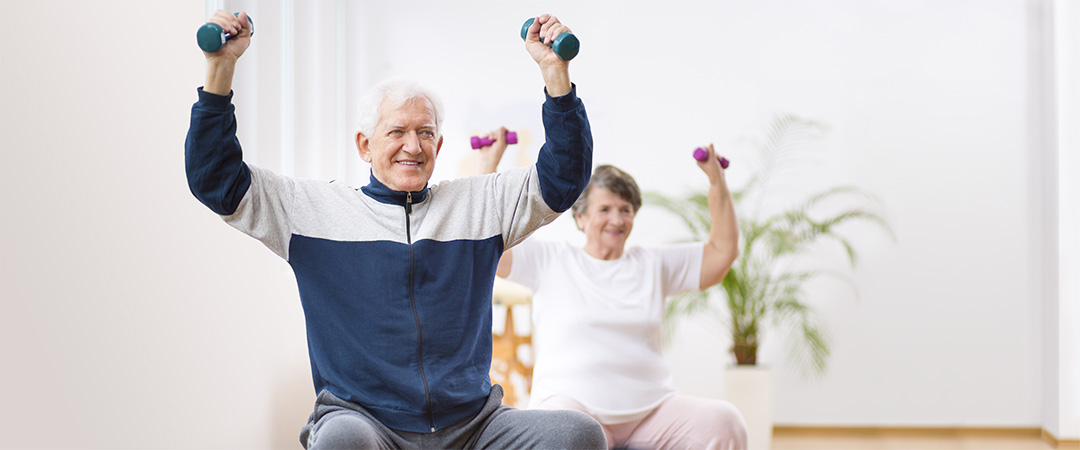 Two seniors in a room lifting light weights on an exercise ball.
