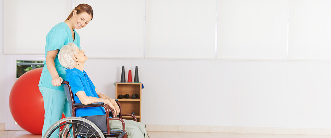 A senior in a wheelchair being wheeled to the rehabilitation room by a nurse.