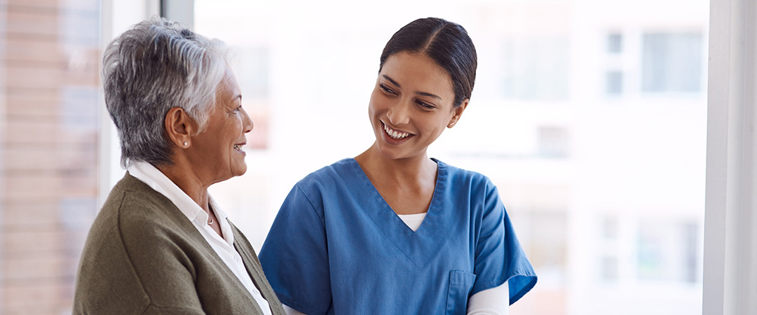 A senior smiling at a nurse who is leading her down a hallway.