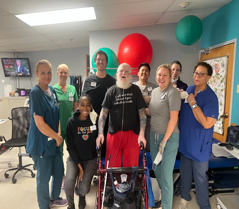 Bearded man with a walker and members of the therapy staff with green and red exercise ball behind them.