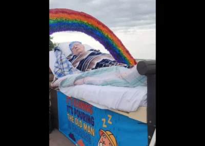 Man on a float with a rainbow over it that is titled It's raining, it's pouring, the old man is snoring.