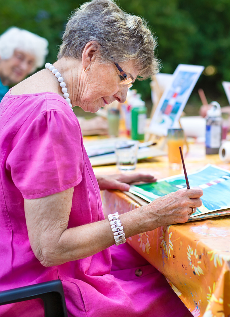 senior resident wearing a pink dress, sitting outside, and painting a picture of a lighthouse