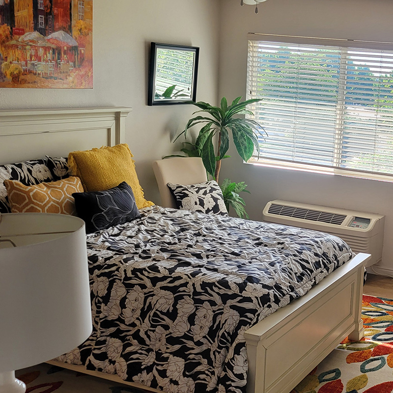 A resident bedroom with a nicely made bed, nightstand, and indoor tree beside a large window.