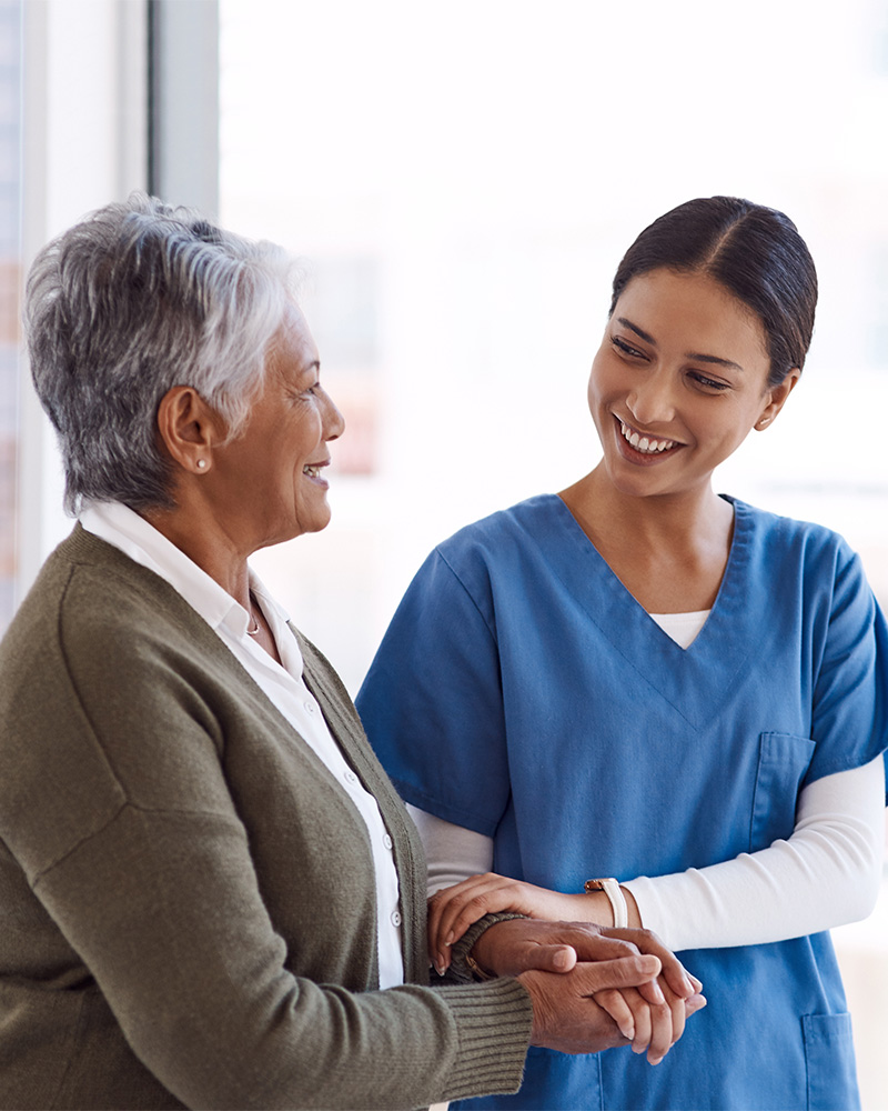 a caregiver holding hands with a mature woman as they walk down a hallway.
