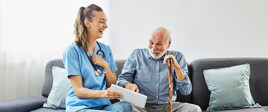 Healthcare worker talking to man on a sofa with a cane, both are laughing.