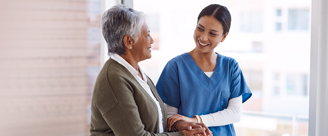 Healthcare worker in blue scrubs walking with senior woman while smiling at each other.