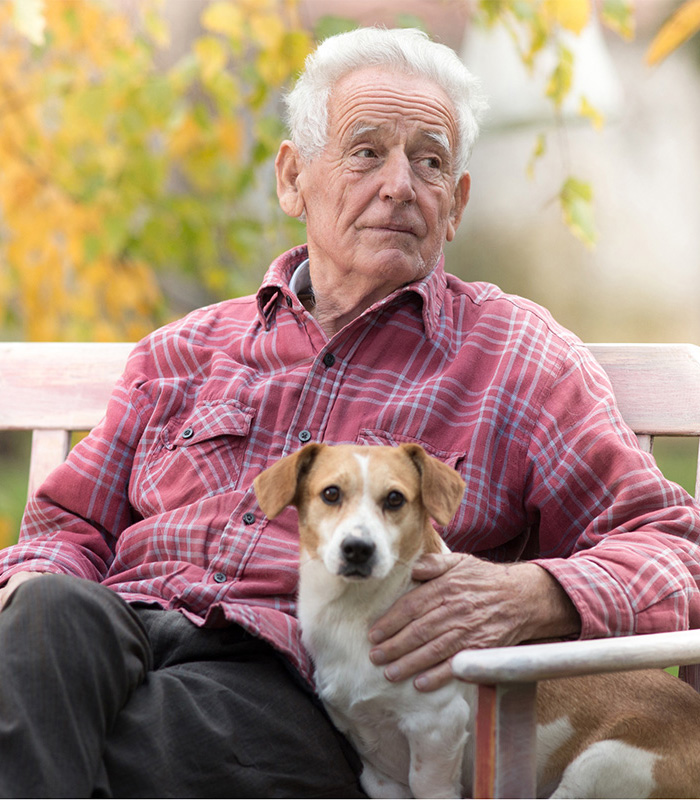 A mature male sitting on a bench outside with his dog beside him.