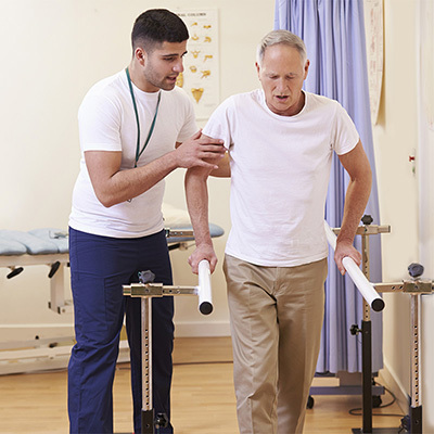 A rehab therapist helping a mature male use the parallel bars safely.