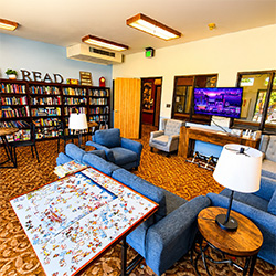 Oakwood Village activity room with puzzle table, blue sofas, television, and large library bookcase full of books.