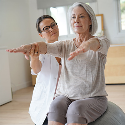 yolo-400x400-94 Therapist helping woman lift her arms while on an exercise ball.